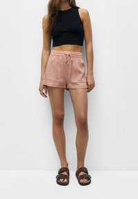 Pantaloni corti rosa in cotone con vita elastica e cordoncino, abbinati a una maglietta nera senza maniche e sandali marroni. Texture liscia.