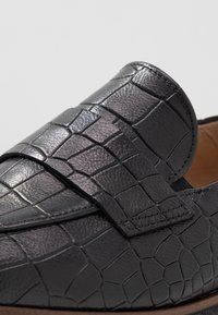 Mocassins en cuir noir avec un motif en crocodile texturé et une fermeture à sangle plate contrastante. Doublure intérieure marron clair.
