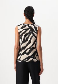 Persona con i capelli intrecciati che indossa un top senza maniche con strisce zebrate nere e beige, abbinato a pantaloni neri solidi, in piedi di spalle.