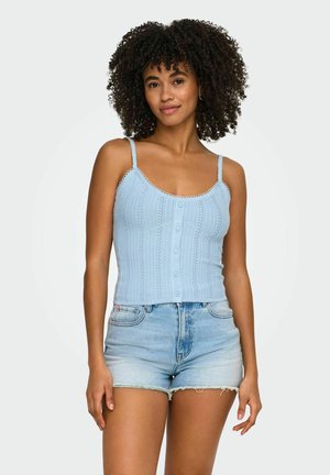Kvinde med krøllet hår iført en lyseblå knap-på strikket tanktop og lyse denim shorts, stående mod en ensfarvet baggrund.