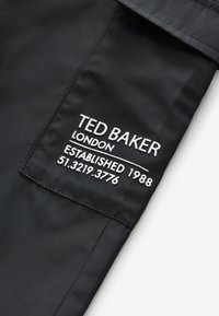 Sort stof med en mat tekstur, der har hvid broderet tekst. Inkluderer logo, etableringsår og geografiske koordinater.