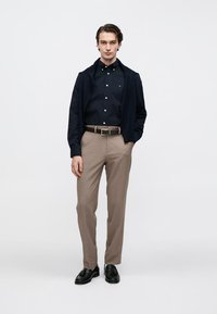 Chemise bleu marine boutonnée sous un cardigan foncé, pantalon kaki et chaussures noires ; présente un logo discret, une coupe ajustée et une texture de tissu lisse.