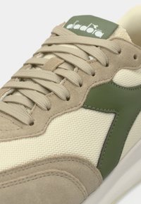 Αθλητικό παπούτσι με μπεζ πλέγμα επάνω μέρος και σουέντ λεπτομέρειες, πράσινο δερμάτινο λογότυπο και υφή ραφών. Τα κορδόνια είναι μπεζ, προσφέροντας ομοιόμορφη εμφάνιση.