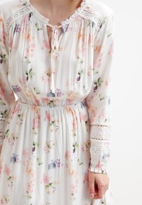 Vestido floral blanco con cuello atado, que presenta patrones en rosa, púrpura y verde. Mangas largas y detalles de encaje en los puños y los hombros.