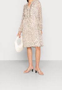 Žena v dlouhých béžových vzorovaných šatech, držící bílou kabelku a obutá v béžových sandálech na podpatku, stojící na šedé podlaze.