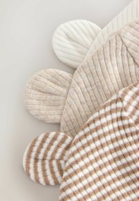 Drie gebreide stoffen hoedsecties in beige en crème, met geribbelde en gestreepte patronen, voorzien van ronde pomponvormen aan de randen.