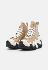 Beżowe, lakierowane wysokie trampki z czarnymi gumowymi podeszwami w kształcie zygzaka, białymi sznurówkami oraz brązowymi akcentami na języku i pięcie.