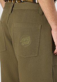 Olive green pants made of textured fabric with a back pocket featuring a circular yellow "Santa Cruz" logo embroidery.