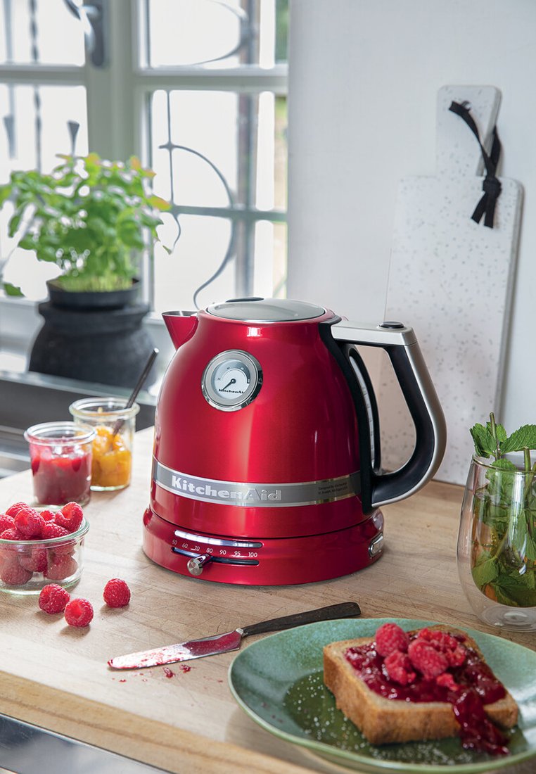 Roter KitchenAid Wasserkocher auf einer Holzküche mit Toast mit Himbeermarmelade, frischen Himbeeren, Marmeladengläsern, Messer und einer grünen Pflanze in einer hellen Küche.