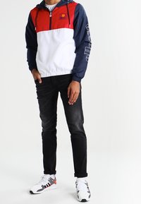 Herrarnas vindjacka i röd, marinblå och vit. Har ett halvmatchande dragkedjestängning, en huva och en elastisk fåll. Kombinerad med svarta jeans och sneakers.