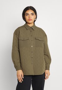 Camisa verde oliva acolchoada com frente de botão, dois bolsos no peito e colarinho. Apresenta um tecido resistente com padrão em losango.