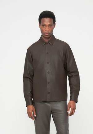 Junger Mann, der ein dunkelbraunes Lederhemd mit Knöpfen und passende Lederhosen trägt, vor einem weißen Hintergrund stehend.