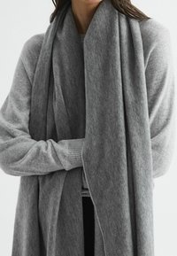En grå uldhalstørklæde hænger om halsen, kendetegnet ved en blød tekstur og et forlænget design, suppleret af et lagdelt gråt outfit nedenunder.