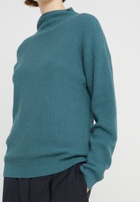 Maglione a collo alto color teal realizzato in tessuto a maglia testurizzata, con maniche lunghe e dettagli a coste all'orlo e ai polsini.