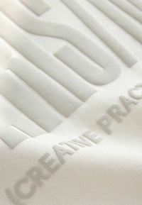 Tissu blanc avec texte en relief "MINIMAL/CREATIVE PRACTICE" dans une finition mate et lisse ; la surface texturée contraste avec le fond plat.