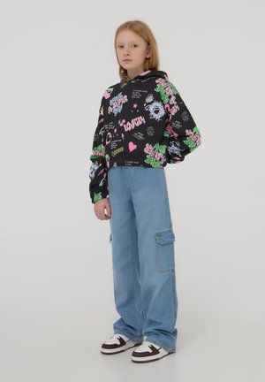 Crop hoodie nera con stampe grafiche colorate; jeans cargo blu con tasche laterali; sneakers bianche con accenti marroni.