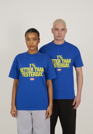 Dwoje dorosłych stojących obok siebie, ubranych w niebieskie koszulki z żółtym napisem „1% lepiej niż wczoraj” na jednolitym jasnym tle.
