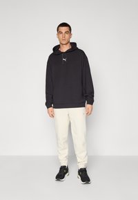 Sudadera negra con cordón y logo en el pecho, combinada con pantalones de chándal de color crema. El modelo lleva zapatillas deportivas negras y está de pie frente a un fondo sencillo.
