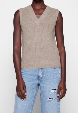 Personne portant un gilet sans manches en maille beige et un jean bleu clair déchiré, debout devant un fond clair uni.