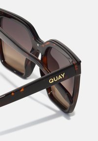 Tortoiseshell sunglasses with dark gradient lenses, thick frames, and gold "QUAY" branding on the side. Smooth plastic texture.