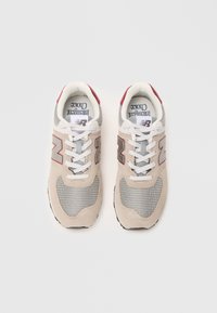 Une paire de sneakers New Balance beige et gris avec des lacets blancs et des accents rouges, vue de dessus sur un fond blanc.