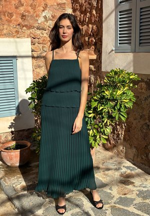 Frau steht im Sonnenlicht vor einer Steinmauer und grünen Pflanzen, trägt ein dunkelgrünes plissiertes Kleid und schwarze Sandalen.