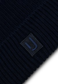Gorro de lana en color azul marino con textura acanalada. Presenta un parche negro cuadrado con un logo estilizado azul. El material parece suave y cálido.