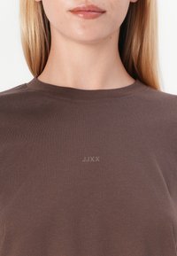 Camiseta de algodão castanha com gola redonda e mangas curtas. Apresenta um discreto logo "JJXX" em relevo no peito. Textura suave.