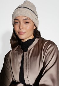 Beanie hat in beige wool blend with a ribbed texture and black horizontal stripe. Worn with a dark, fitted sweater and satin bomber jacket.