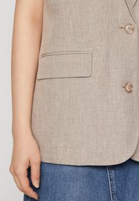 Beige Leinenblazer mit einer einzelnen Brusttasche, zwei Knöpfen und einer strukturierten Passform. Die Textur ist leicht strukturiert und leicht.