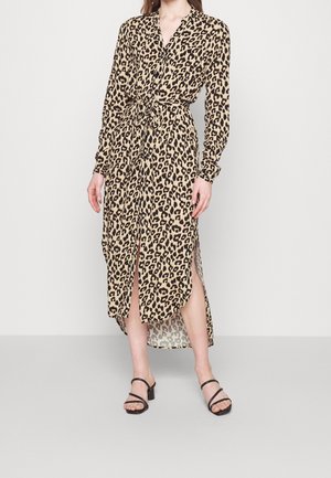 Kvinde iført en beige og sort leopardmønstret midikjole med lange ærmer, bindebånd i taljen og frontslids, kombineret med sorte sandaler med remme.