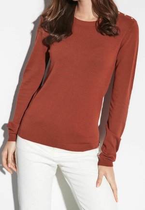 Kvinde iført en tætsiddende rustfarvet langærmet sweater med knapper på skulderen, kombineret med hvide bukser, stående mod en hvid baggrund.
