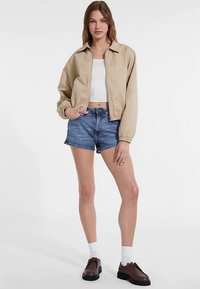 Giacca corta beige con polsini elasticizzati, indossata sopra una canotta bianca e pantaloncini di denim blu. Scarpe stringate marroni completano il look.