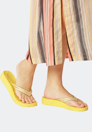 Feet wearing beige flip-flops with thick textured yellow soles, paired with pants featuring vertical stripes in orange, yellow, black, and beige.