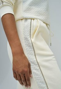 Gros plan sur une main reposant sur un pantalon de jogging crème avec une rayure texturée et un taille élastique à cordon, associé à un haut crème texturé assorti.