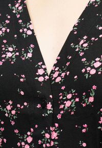 Μαύρο μπλούζα με λαιμόκοψη σε σχήμα V, διακοσμημένη με μικρά ροζ ρόδα και πράσινα φύλλα, κλειστή με ένα μόνο μαύρο κουμπί.