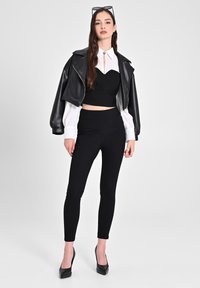 Giacca in pelle nera corta sopra una maglia nera aderente con scollatura a forma di cuore, abbinata a pantaloni neri a vita alta e tacchi neri.