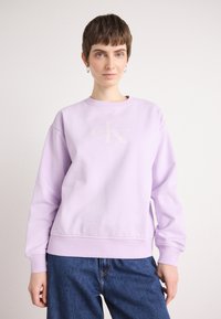 Lilla genser laget av bomullsblanding, med rund hals og ribbestrikkede ermer. Framsiden har et preget "Calvin Klein"-logo.