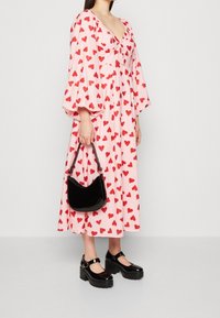 Kvinna som bär en rosa midiklänning med röda hjärtan, svarta platå-Mary Jane-skor och håller en liten svart handväska.