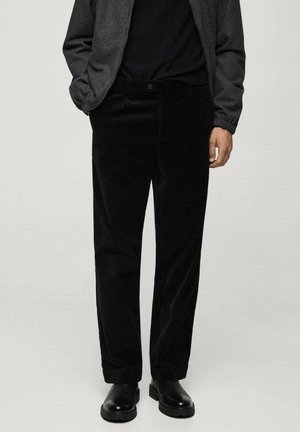 Mann, der schwarze Cordhosen, schwarzes Hemd, dunkelgraue Jacke und schwarze Lederschuhe trägt, steht vor hellem Hintergrund.