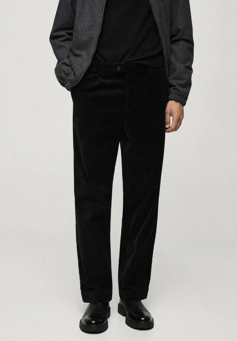 Homme portant un pantalon en velours côtelé noir, une chemise noire, une veste gris foncé et des chaussures en cuir noires, se tenant devant un fond clair.