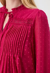 Blusa leggera fucsia con scollatura a rouches, pieghe e motivi floreali ricamati. Tessuto trasparente con dettagli in pizzo intricati.
