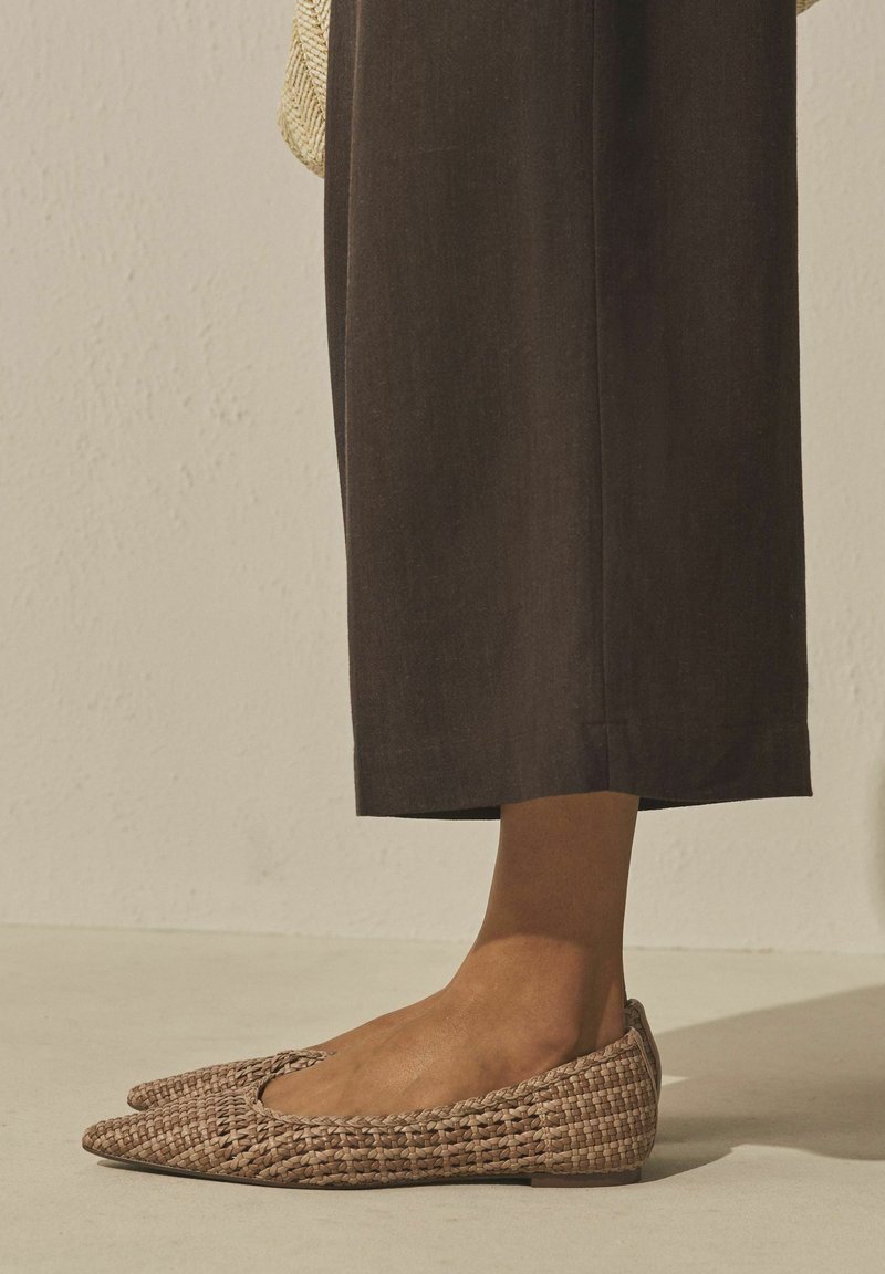 Flettede lysebrune flade sko båret med mørke korte bukser, stående på et lyst glat gulv mod en lys, tekstureret væg.