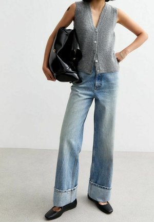 Vrouw draagt een grijze mouwloze, dichtgeknoopte vest, wijd uitlopende opgerolde spijkerbroek, zwarte platte schoenen, en houdt een grote zwarte leren tas onder haar arm.