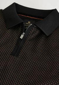 Polo noir avec un col texturé et une patte zippée, orné d'un motif géométrique dans des tons de brun foncé et clair. Étiquette de marque visible à l'intérieur.