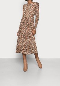 Brun blomsterkjole med lange ærmer, en rund udskæring og en plisseret nederdel. Kombination med beige ankelstøvler på en neutral baggrund.