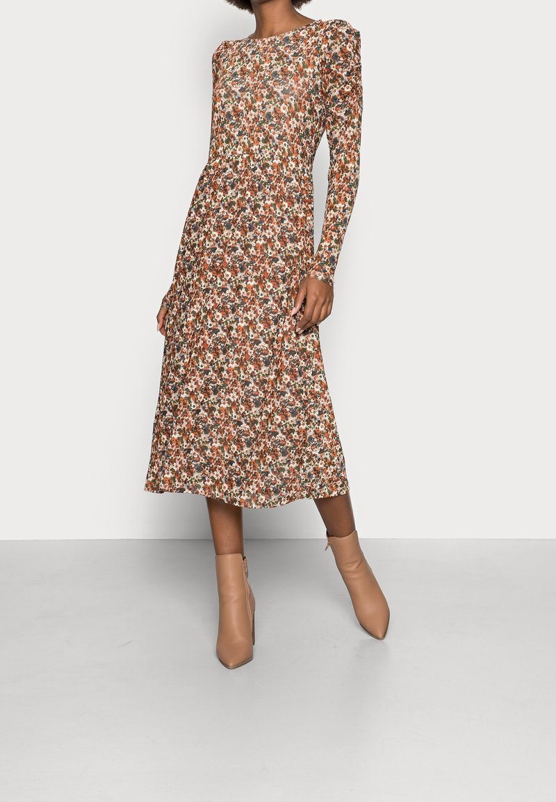 Brun blomsterkjole med lange ærmer, en rund udskæring og en plisseret nederdel. Kombination med beige ankelstøvler på en neutral baggrund.