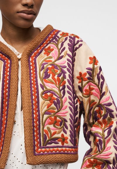 Gros plan d'une personne portant une veste crème avec des bordures marron, brodée de motifs floraux et de vignes en violet, orange, rose et vert.
