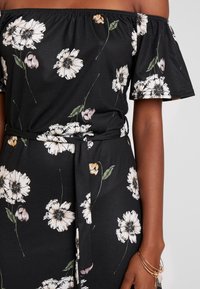 Černé šaty s odhalenými rameny a květinovým vzorem v bílé a růžové barvě. Mají elastický výstřih, krátké rukávy a opasek na zavázání v pase.