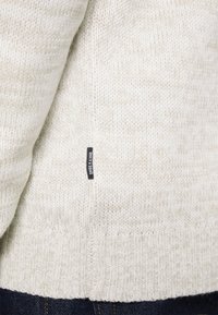 Sweter w kolorze kremowym, wykonany na drutach, noszony na ciemnoniebieskich dżinsach, z małą czarną metką z białym napisem na bocznej szwie.